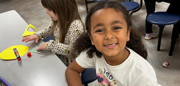 girl sitting at table smiling with another girl working on craft project