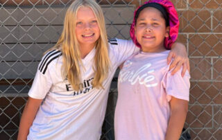 a girl with arm around another girl in front of chain link fence