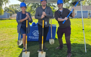 3 men in blue hardhats with shovels on green grass lawn